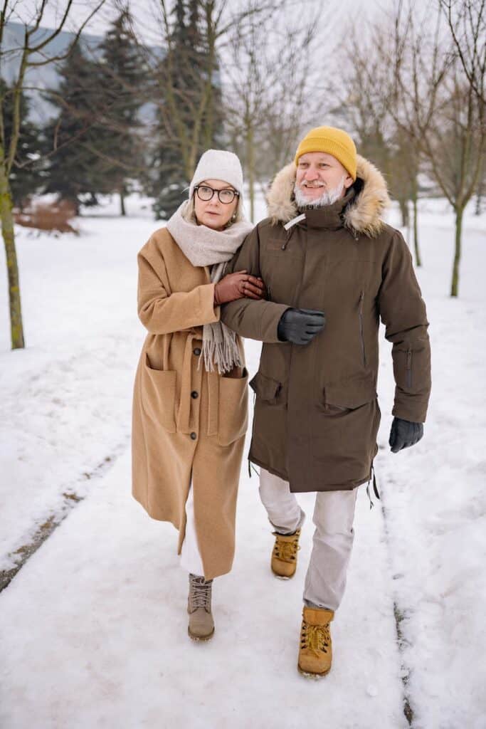 an elderly couple walking on snow covered ground holding arms.